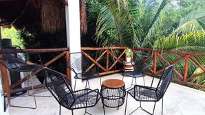 Un espace extérieur abrité, aménagé avec des chaises en métal et une table en bois, entouré d’une balustrade en bois et d’une végétation luxuriante.