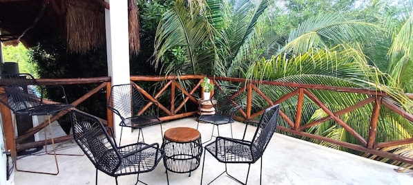 A covered outdoor seating area with metal chairs and a wooden table, surrounded by a wooden railing and lush greenery.
