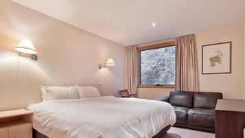 A bedroom with a bed, bedside tables, a chair, a desk, a window with curtains, and framed botanical prints on the wall.