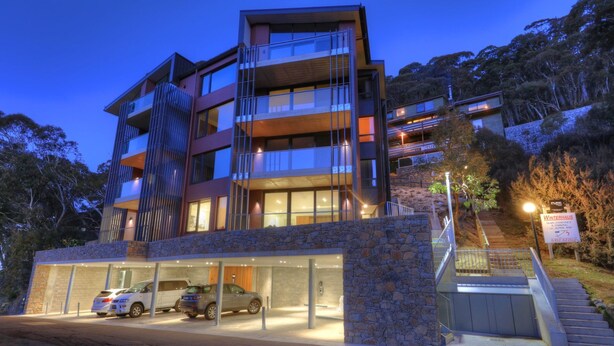 A multi-story hotel with a stone facade, multiple balconies, and a parking area with several cars.