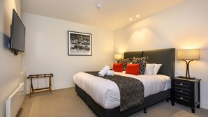 A hotel room with a bed, a bedside table with a lamp, a small wooden stool, a wall-mounted TV, and a framed picture on the wall.