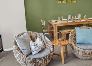 Un coin repas avec des chaises en osier, une table en bois dressée pour quatre personnes et un mur vert orné d’œuvres d’art.