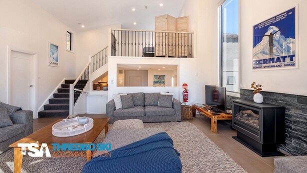 A spacious living area with a staircase leading to a loft, a fireplace, and a large window with a poster. - Blues Air 4 - Woodridge (Woodridge)
