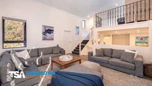 A spacious living area with two grey sofas, a coffee table, and a large window with a view of trees. A staircase leads to an upper level with a balcony. - Blues Air 4 - Woodridge (Woodridge)