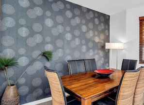 Un coin repas avec une table en bois, des chaises en osier, un vase avec une plante et un mur orné d’un motif circulaire.