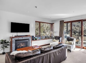A modern living room with a fireplace, a flat-screen TV, a sofa, and a balcony with a view of trees. - Elevation 5 - Central Village (Central Village)