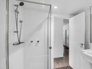 A modern bathroom with a glass shower enclosure, a white sink, and a door leading to another room. - Elevation 5 - Central Village (Central Village)