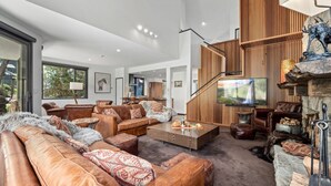 Een ruime woonkamer met leren banken, een bijzettafeltje en een open haard. Er is een trap met houten lambrisering en een aan de muur bevestigde televisie.