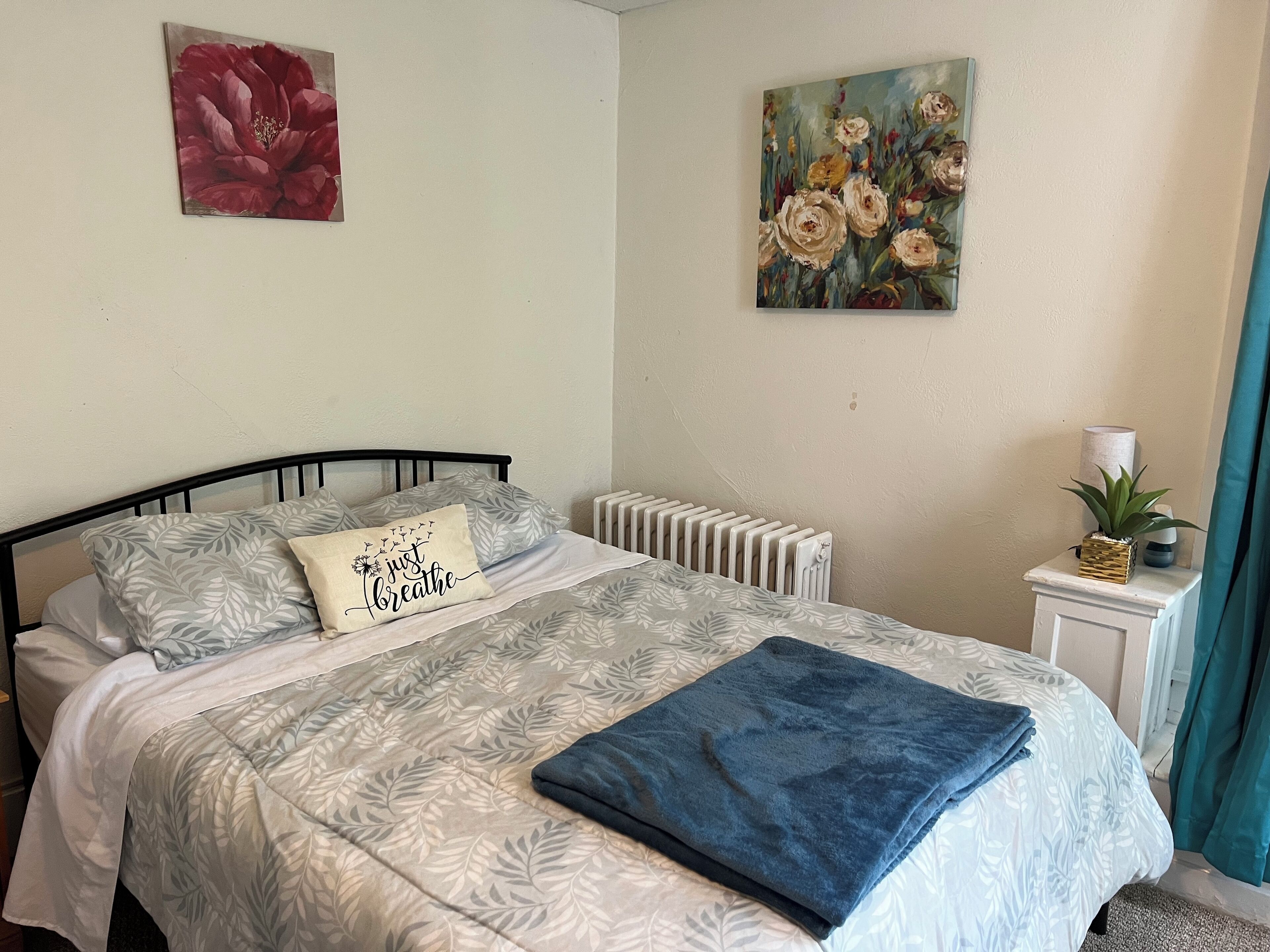 Una cama bien hecha con ropa de cama estampada, un cojín con una frase motivacional y dos cuadros florales en las paredes.