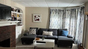 Un salon comprenant un canapé, une table basse et une cheminée. On y trouve également un téléviseur posé sur un meuble et une bibliothèque agrémentée d’objets décoratifs.