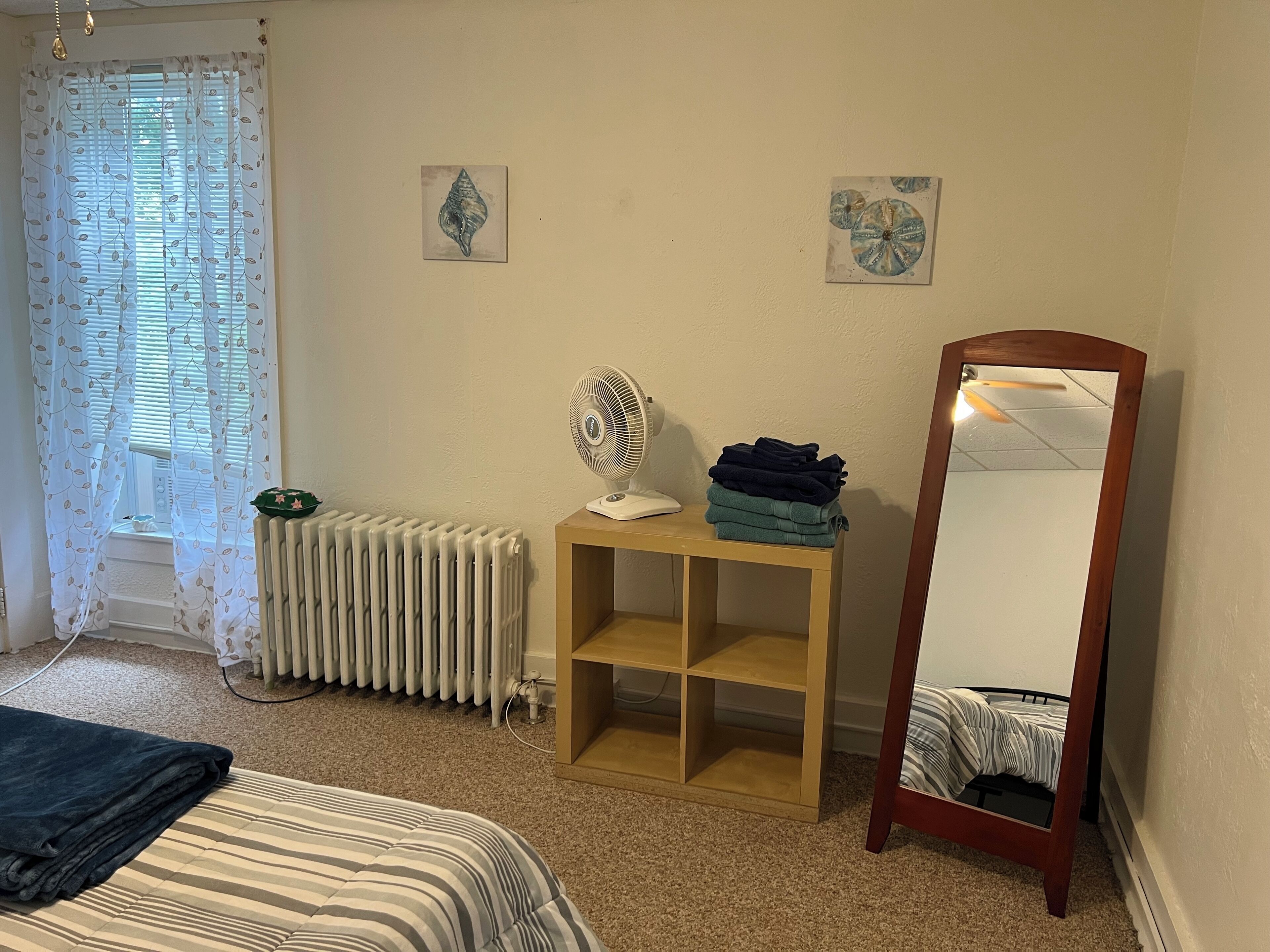 A bedroom with a bed, a radiator, a standing fan, a mirror, and a shelf with folded towels.