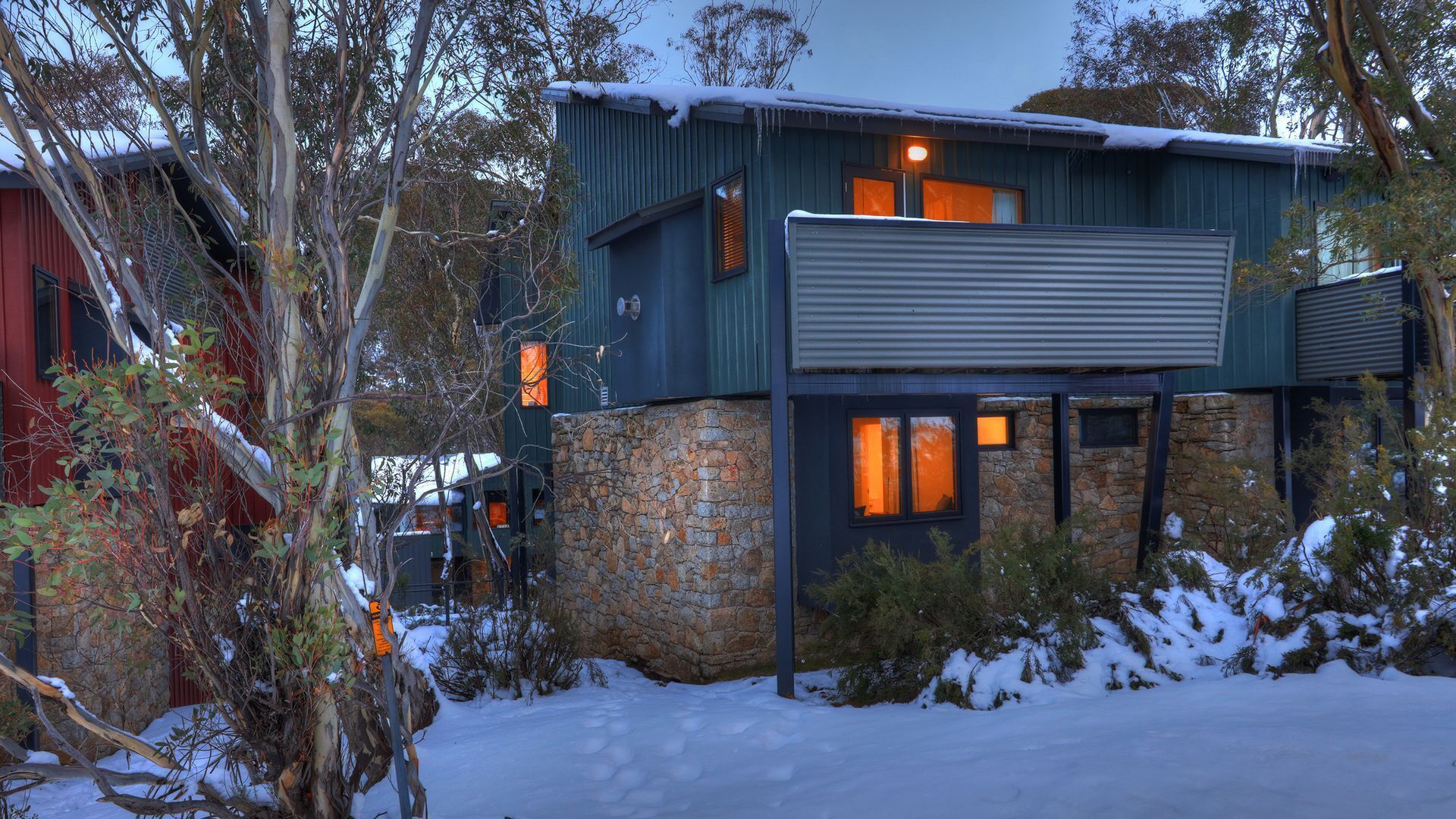 A modern house with a stone foundation and a metal roof, surrounded by snow-covered ground and trees.