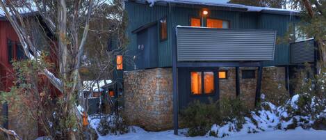 A modern house with a stone foundation and a metal roof, surrounded by snow-covered ground and trees.