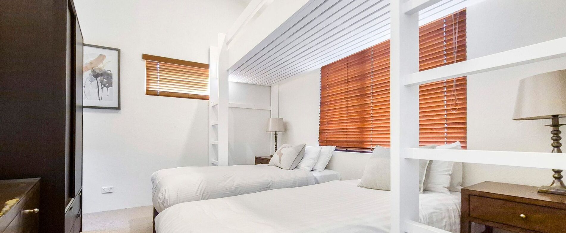 A bedroom with two beds, wooden blinds, a bedside table with a lamp, and a framed picture on the wall.