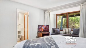 Ein modernes Schlafzimmer mit Bett, Fernseher, Fenster mit Vorhängen und Blick ins Grüne.