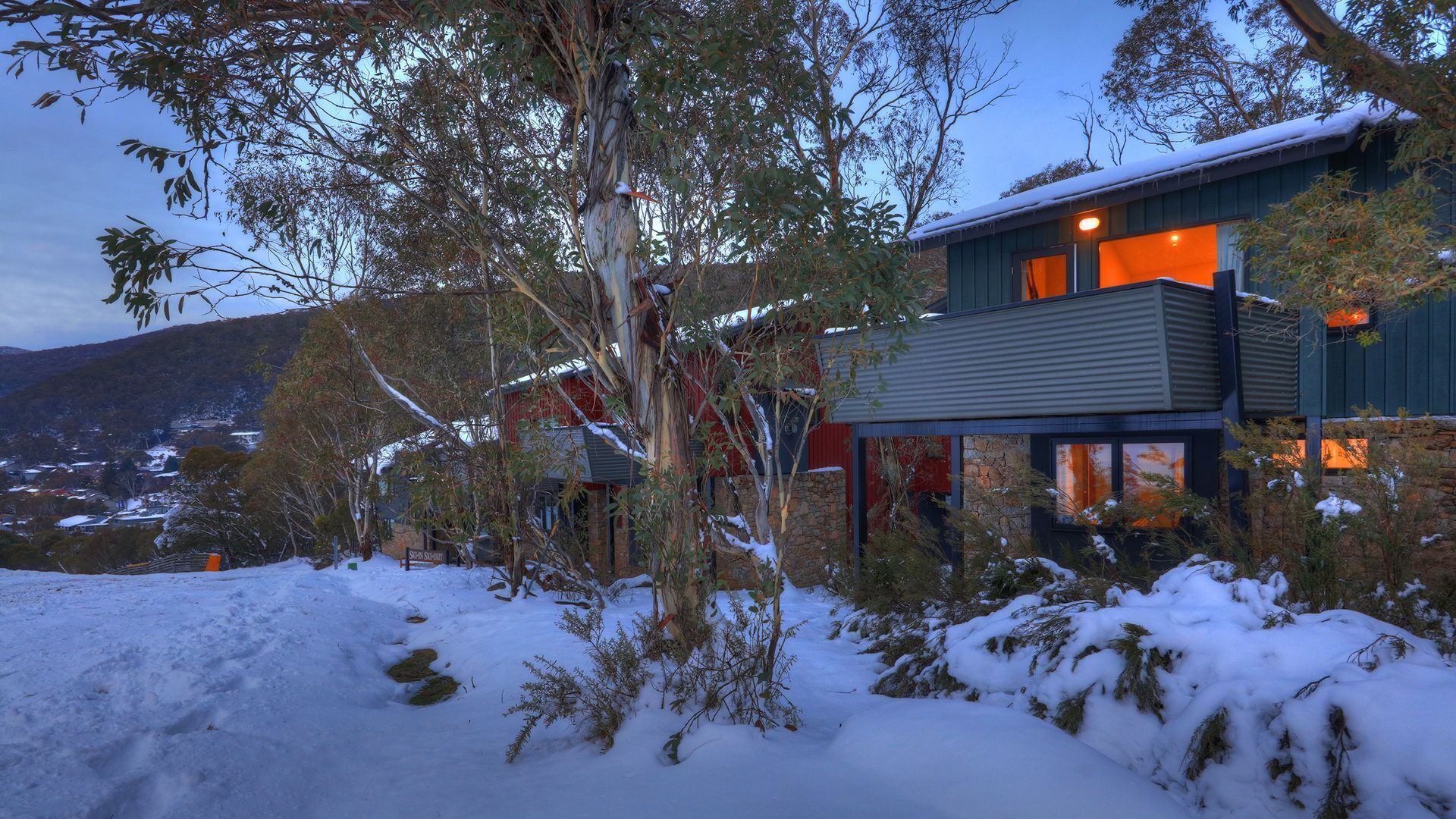 A modern two-story house with a stone foundation, surrounded by snow-covered ground and trees.