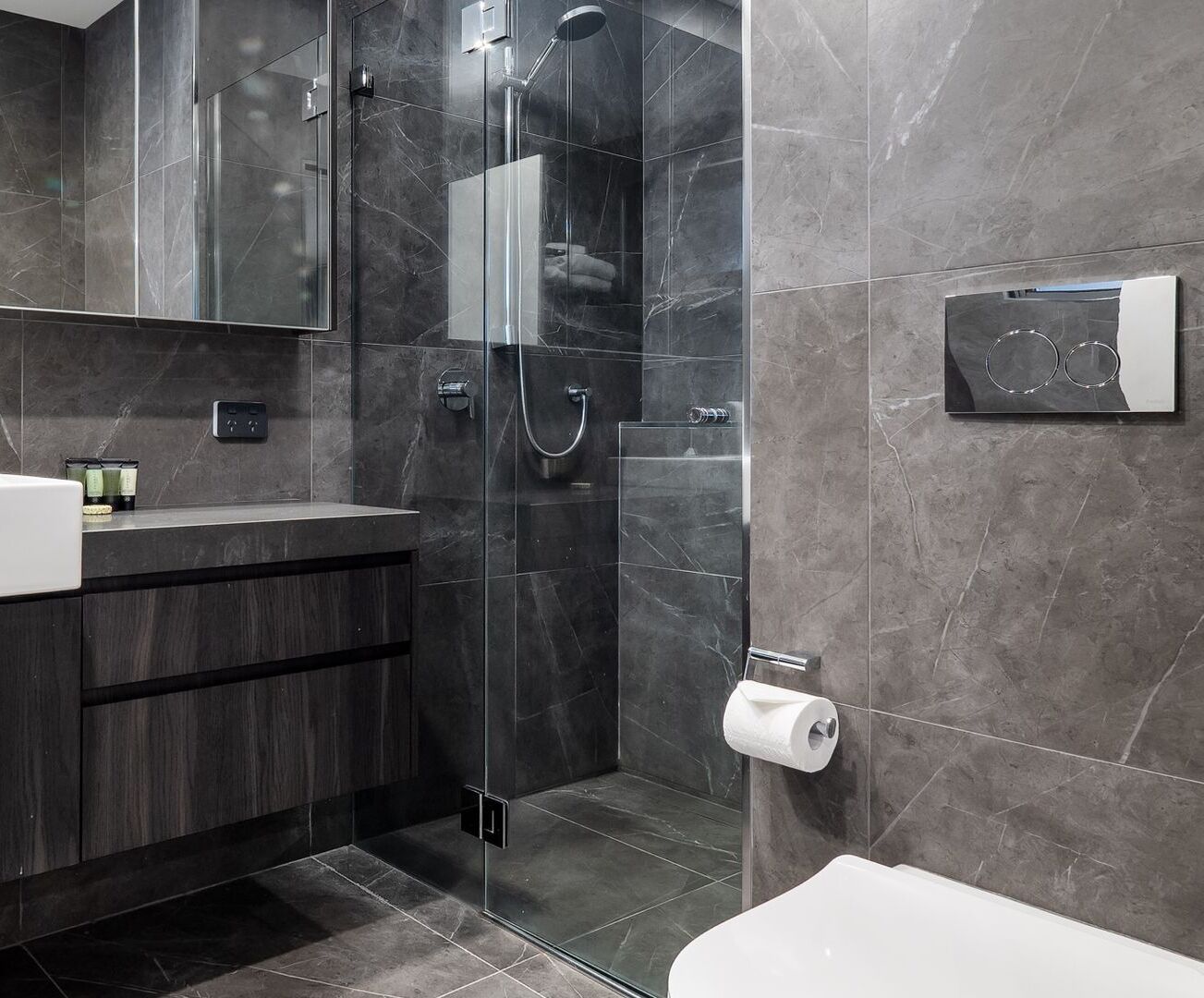 Ein modernes Badezimmer mit einer Duschkabine, dunklen Marmorfliesen, einem Holzwaschtisch und einem Toilettenpapierhalter.