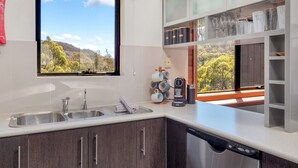 A modern kitchen with a stainless steel sink, a coffee machine, and a dishwasher. There is a window with a view of trees and hills.