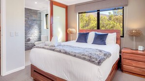 A modern bedroom with a large bed, a stone accent wall, a window with a view of trees, and a wooden nightstand with a lamp.