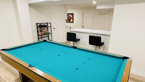 A pool table in a room with a wooden floor, white walls, and a small bar area with two black chairs. - Modern Thorold Apt w/ Pool Table (Thorold)