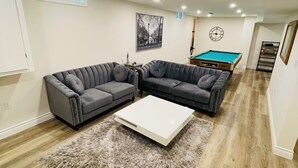 A modern living room with a sectional sofa, a coffee table, a pool table, and a framed cityscape picture on the wall. - Modern Thorold Apt w/ Pool Table (Thorold)