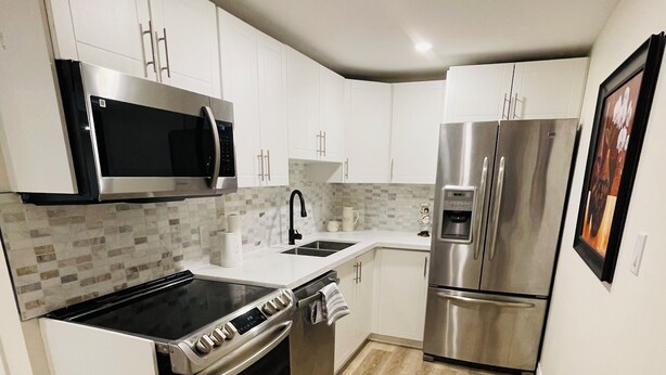 A modern kitchen with white cabinetry, stainless steel appliances, a tiled backsplash, and a wooden floor. - Modern Thorold Apt w/ Pool Table (Thorold)