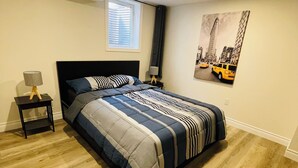A bedroom with a bed, a nightstand, a lamp, a window with blinds, and a cityscape painting on the wall. - Modern Thorold Apt w/ Pool Table (Thorold)