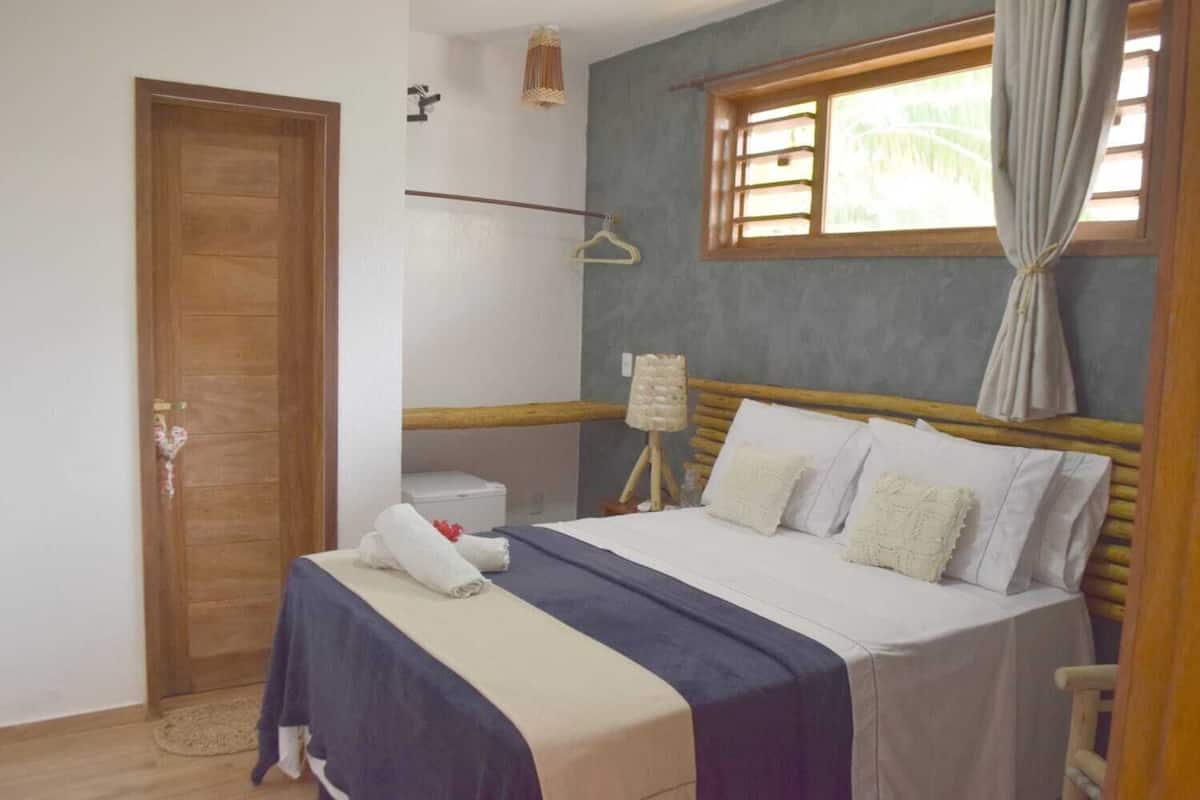 Ein Schlafzimmer mit einem hölzernen Bett, einem Fenster mit Vorhängen, einem Nachttisch und einer Pendelleuchte.