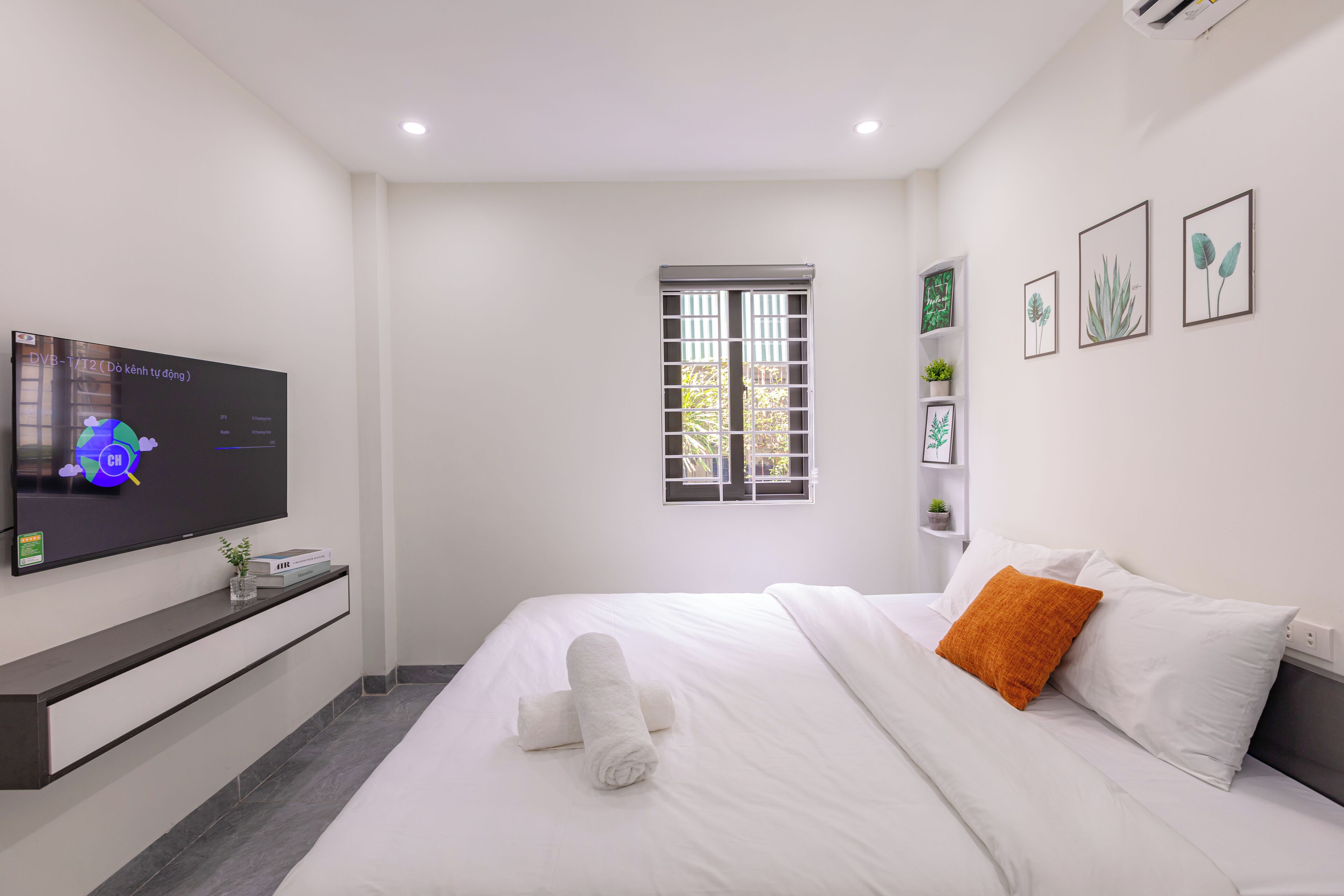 A modern hotel room with a large bed, a flat-screen TV mounted on the wall, a window with a view of greenery, and a small shelf with decorative items.