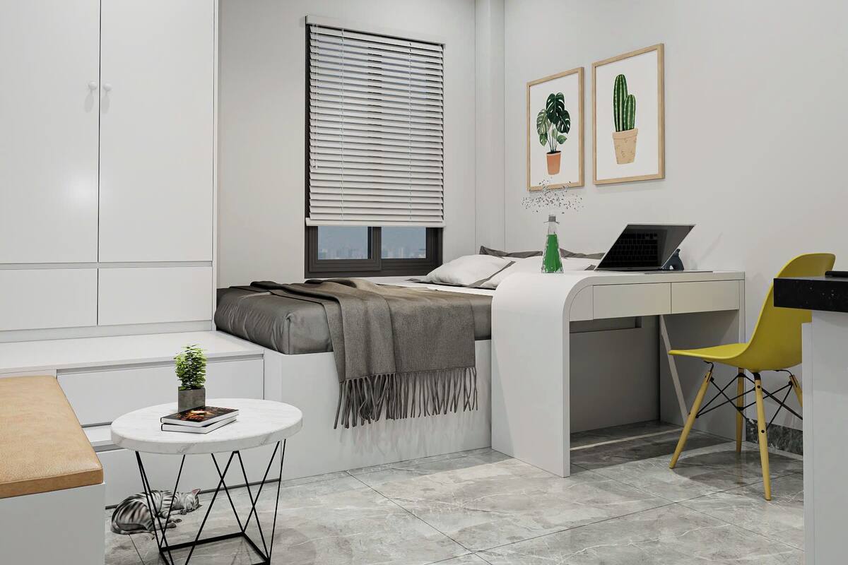 Ein schlichtes, modernes Schlafzimmer mit Bett, Schreibtisch und Stuhl, einem kleinen runden Tisch mit Pflanze und einem Fenster mit Jalousien.