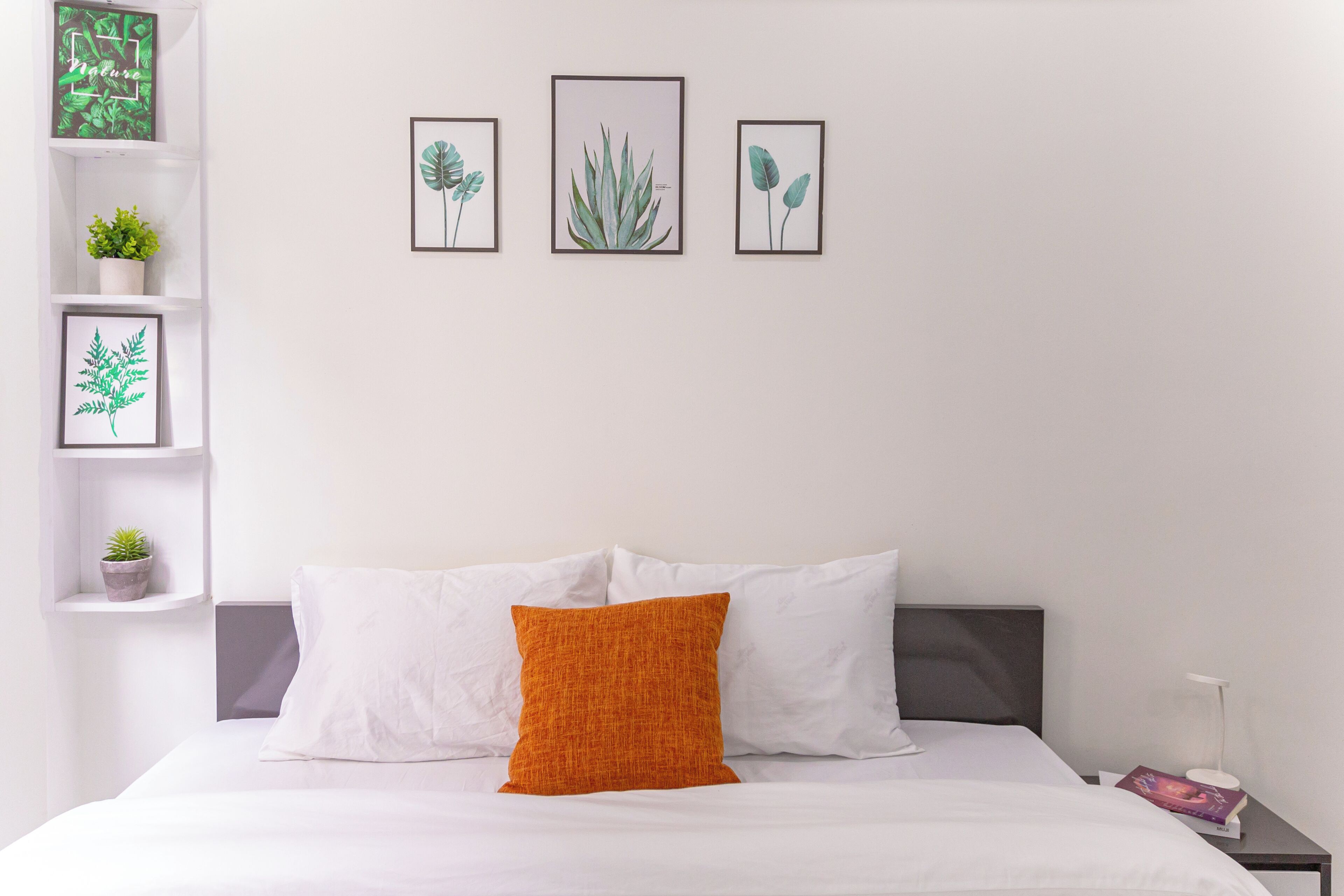 A neatly made bed with white and orange pillows, a grey headboard, and a small bedside table with a lamp and book. There are three framed botanical prints on the wall above the bed.