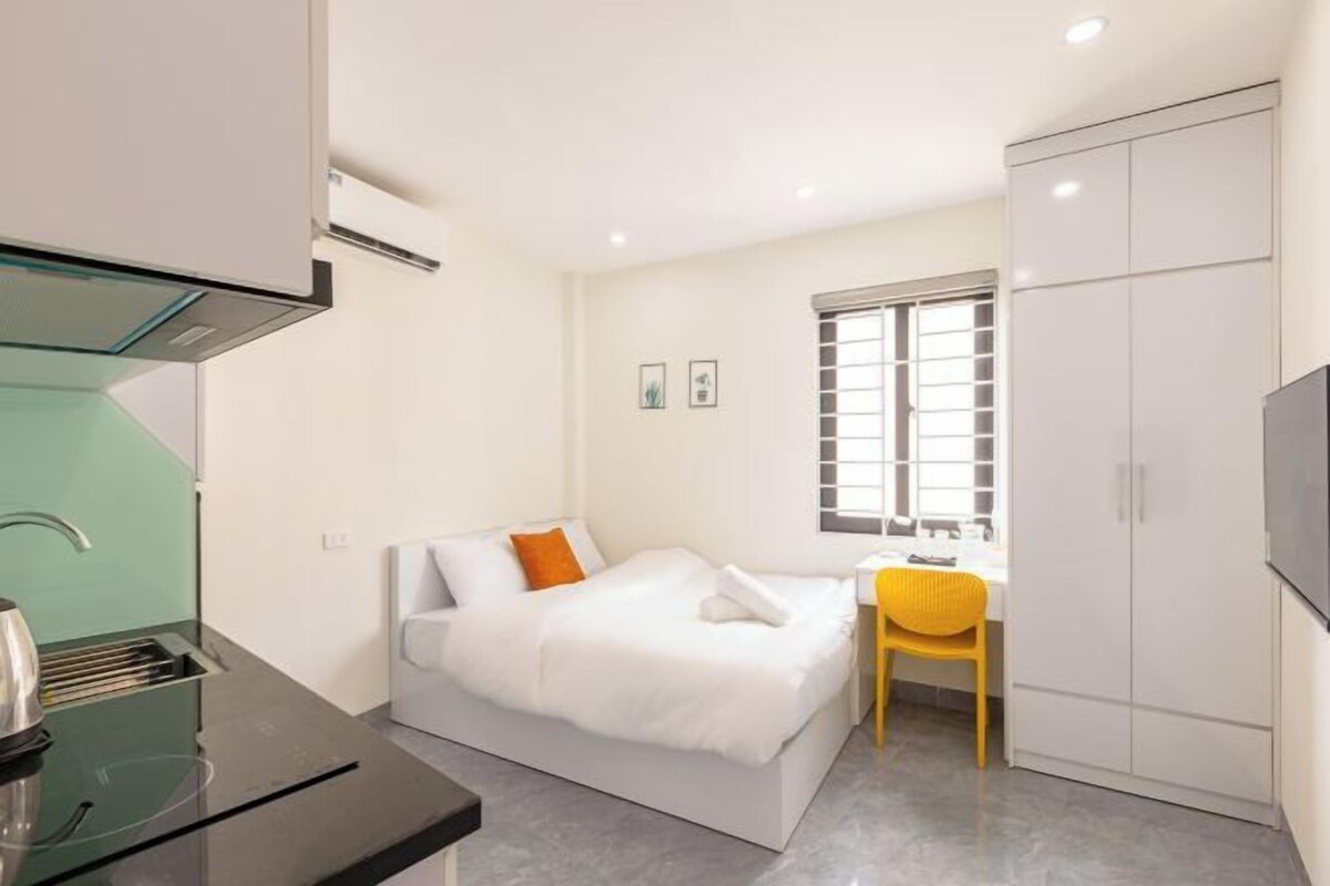 A compact hotel room with a bed, a small desk, a chair, a wardrobe, and a kitchenette.