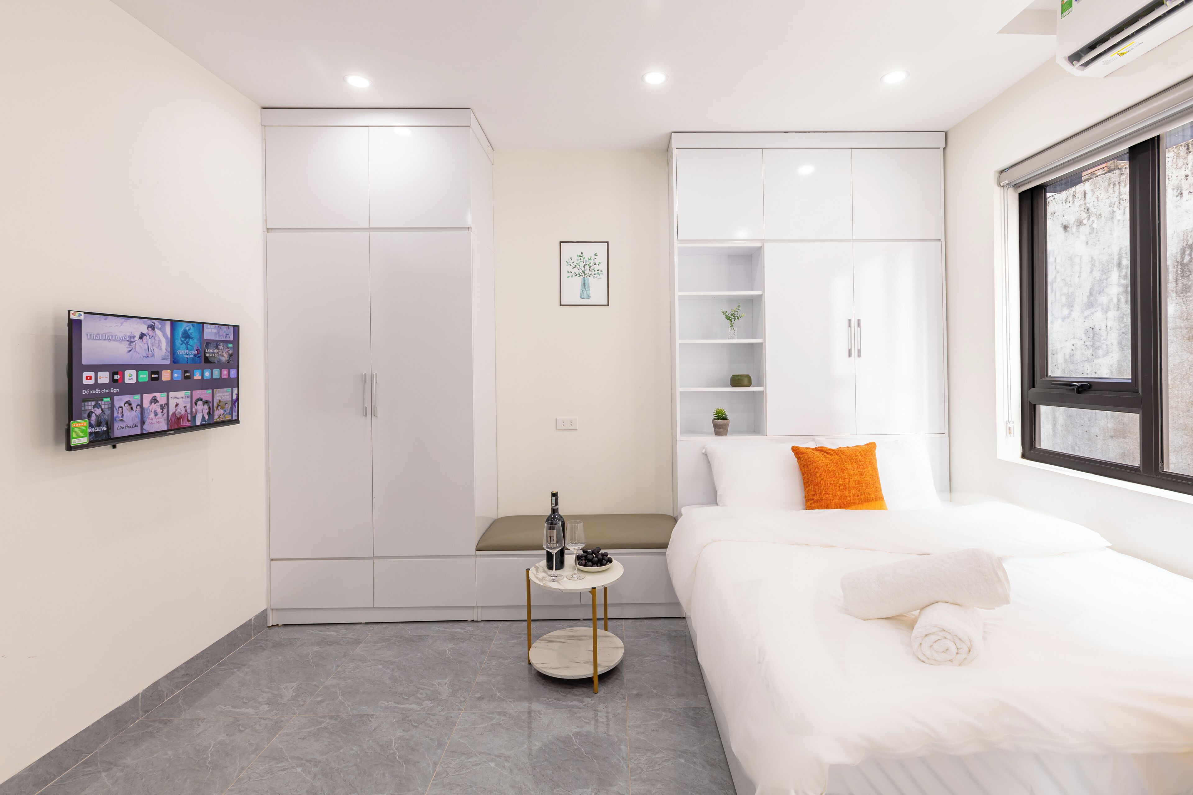 Una habitación de hotel moderna con una cama, una mesita con una botella y vasos, un televisor en la pared y un armario.