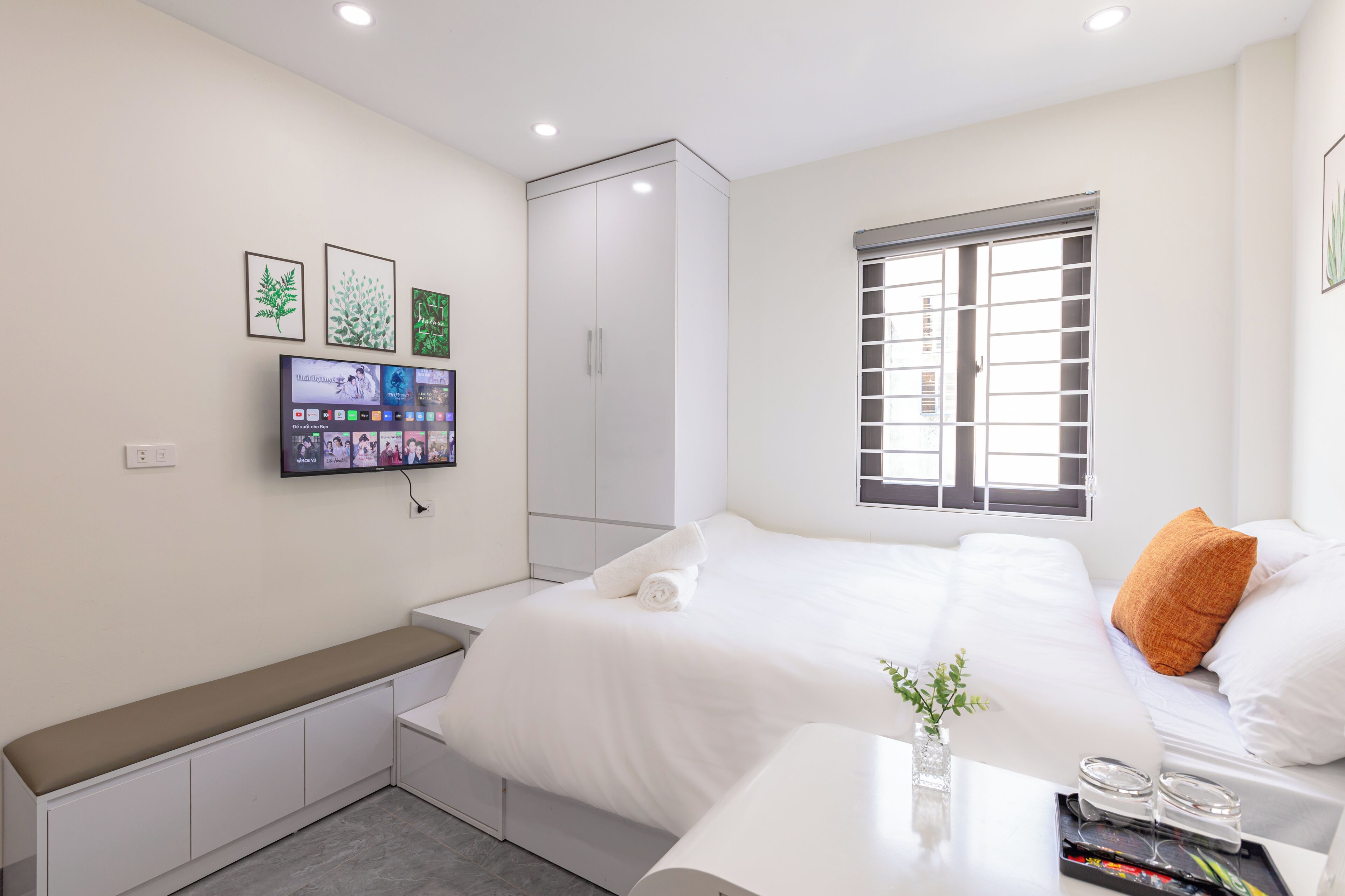 A modern hotel room with a bed, a bench, a wardrobe, a TV, and a window with blinds.