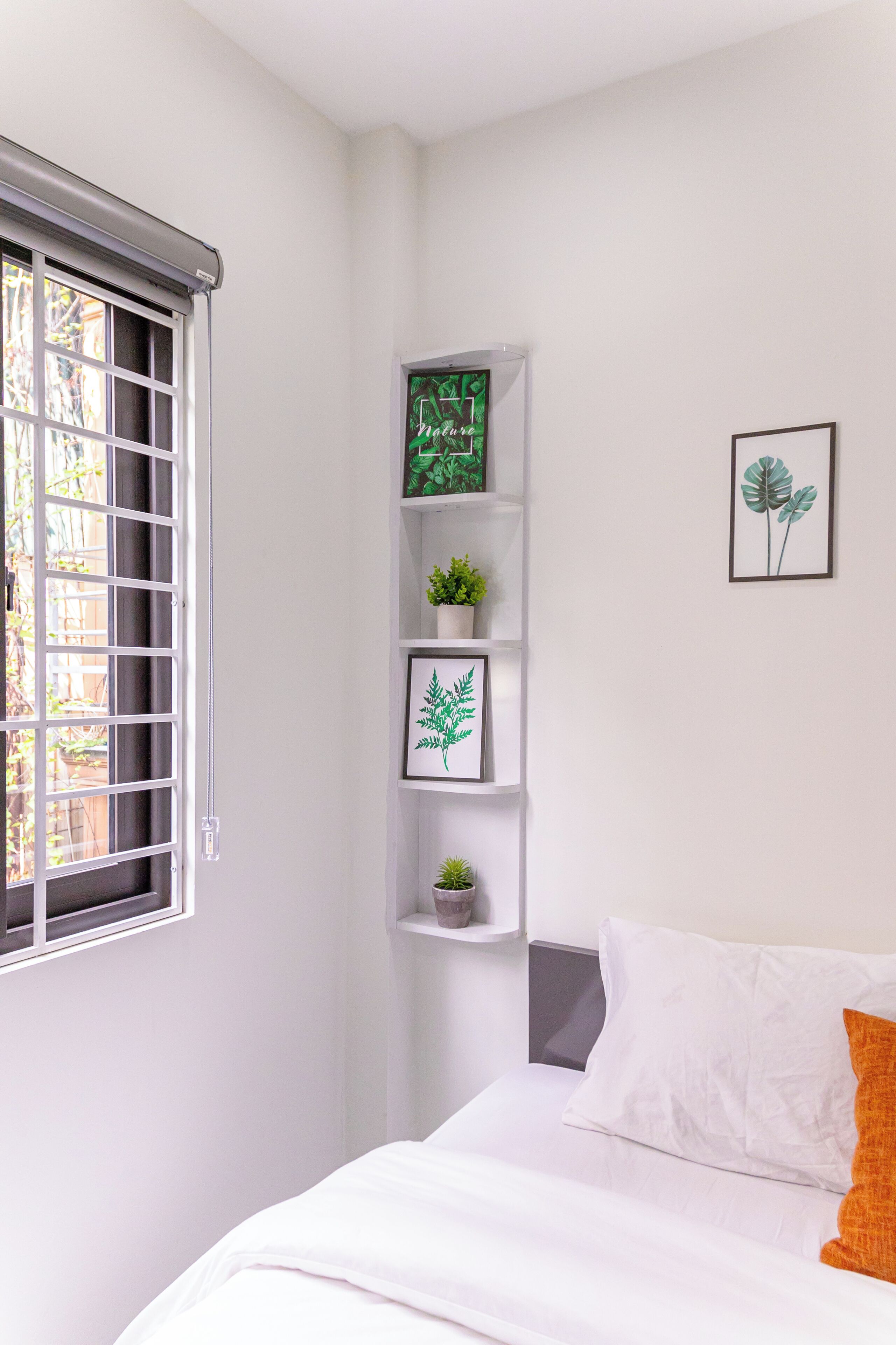 Ein Schlafzimmer mit einem Bett, einem Fenster mit Jalousien, einem Regal mit Pflanzen und gerahmten Bildern sowie einer Wand mit einer gerahmten Pflanze.