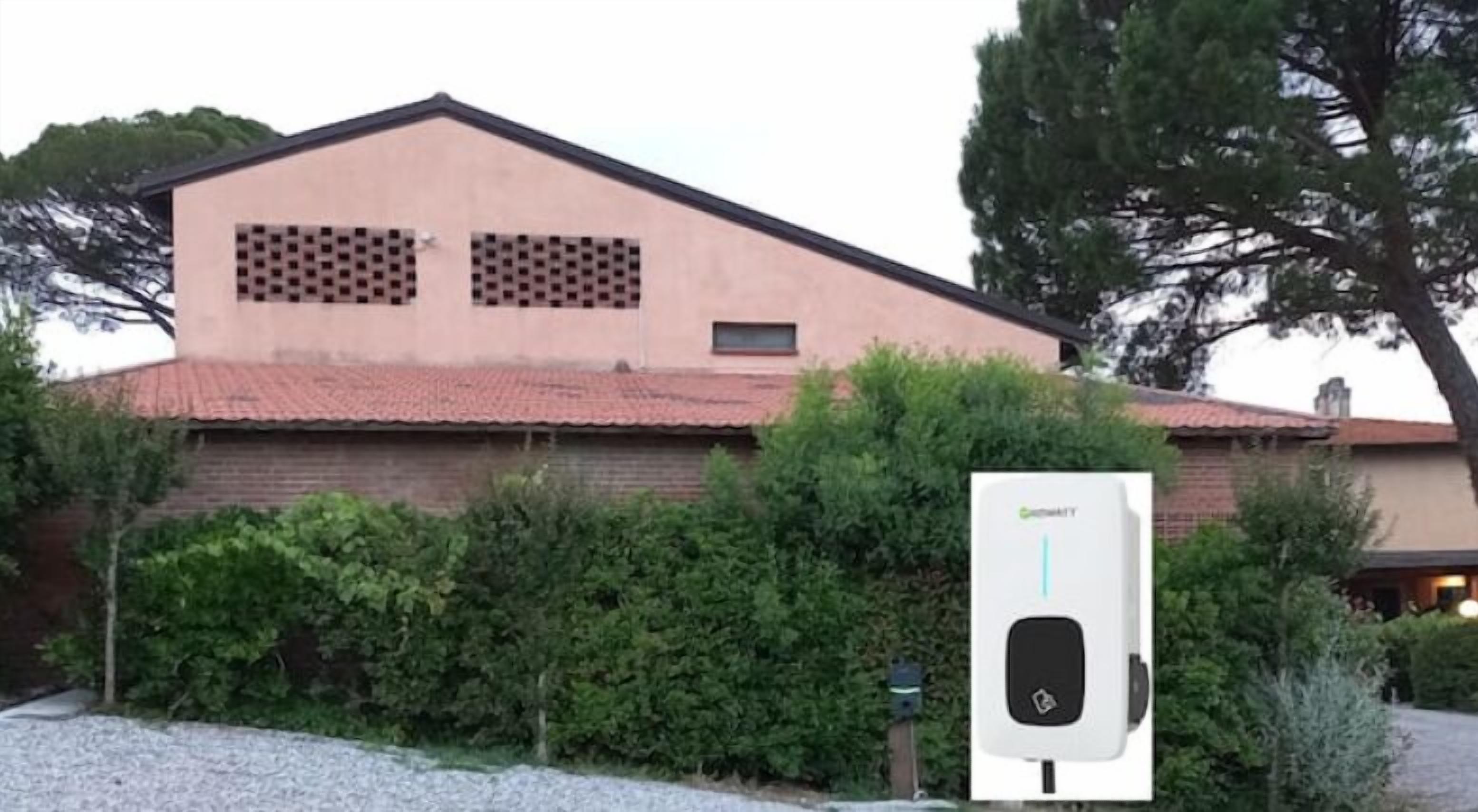 Un edificio con fachada de ladrillo y techo de tejas, un sistema de paneles solares al frente y un árbol a la derecha.