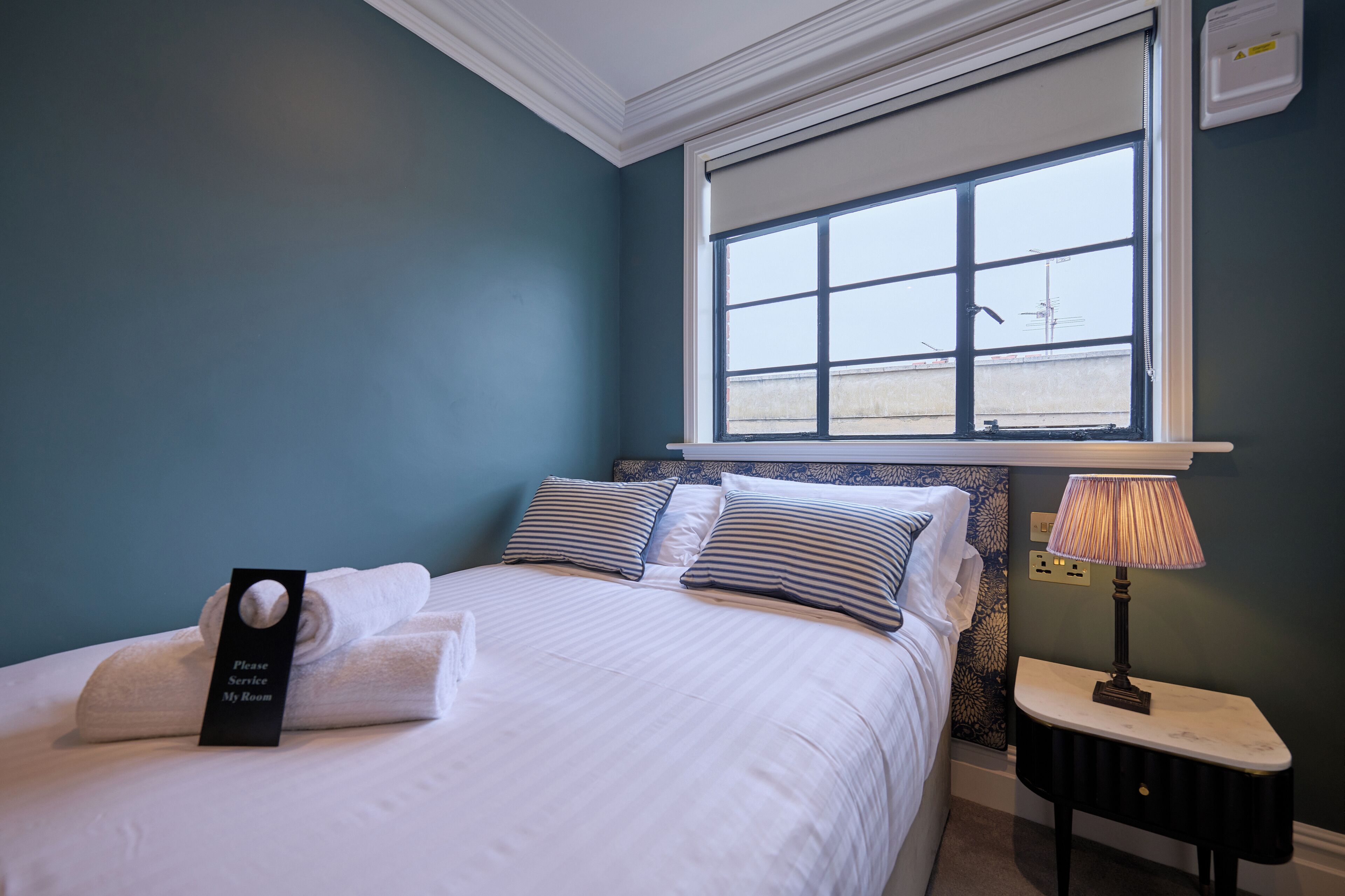 A neatly made bed with a sign indicating the room, a bedside table with a lamp, and a window with a view of the outside.