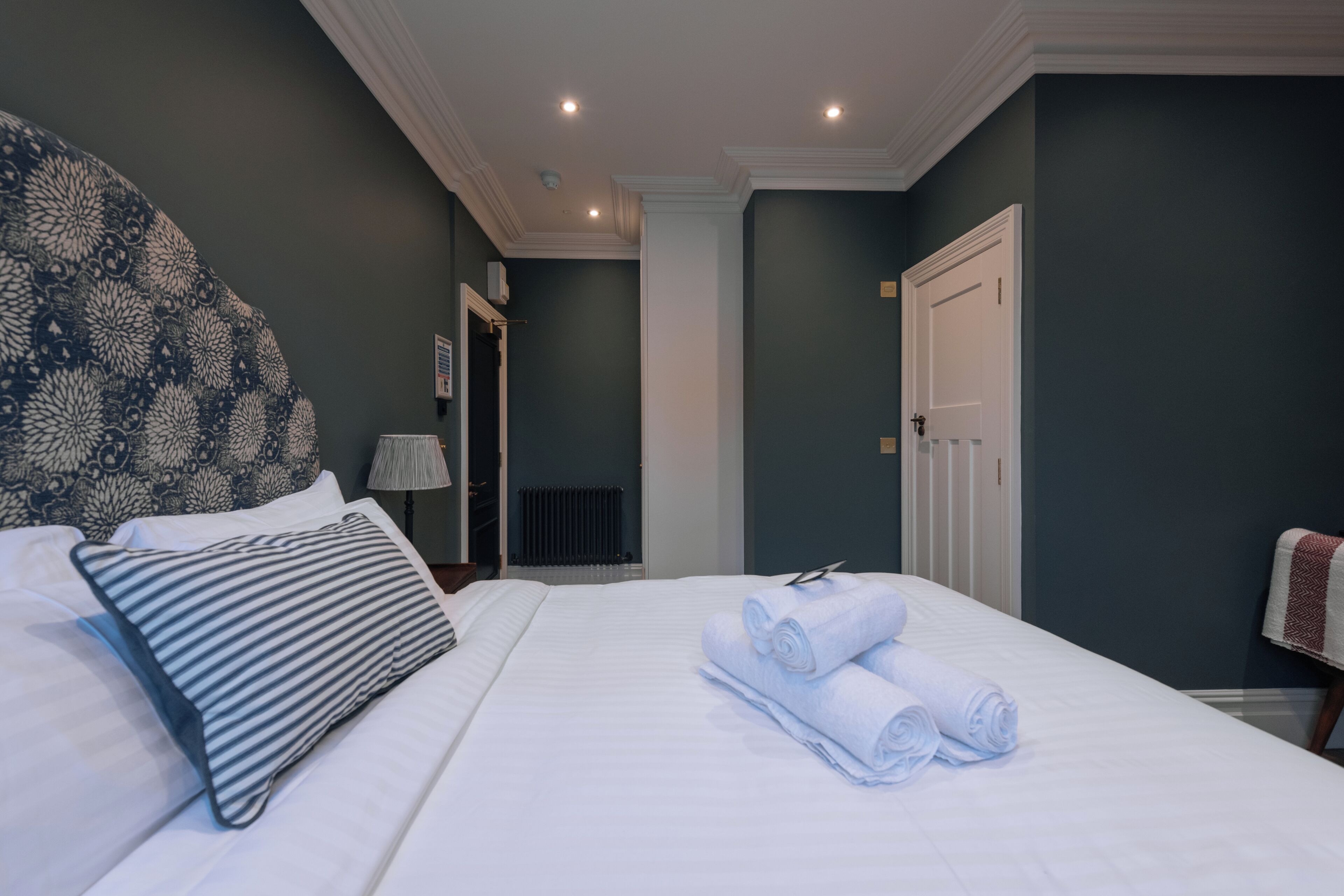A hotel room with a large bed, a headboard with a floral pattern, and a neatly made bed with rolled towels.