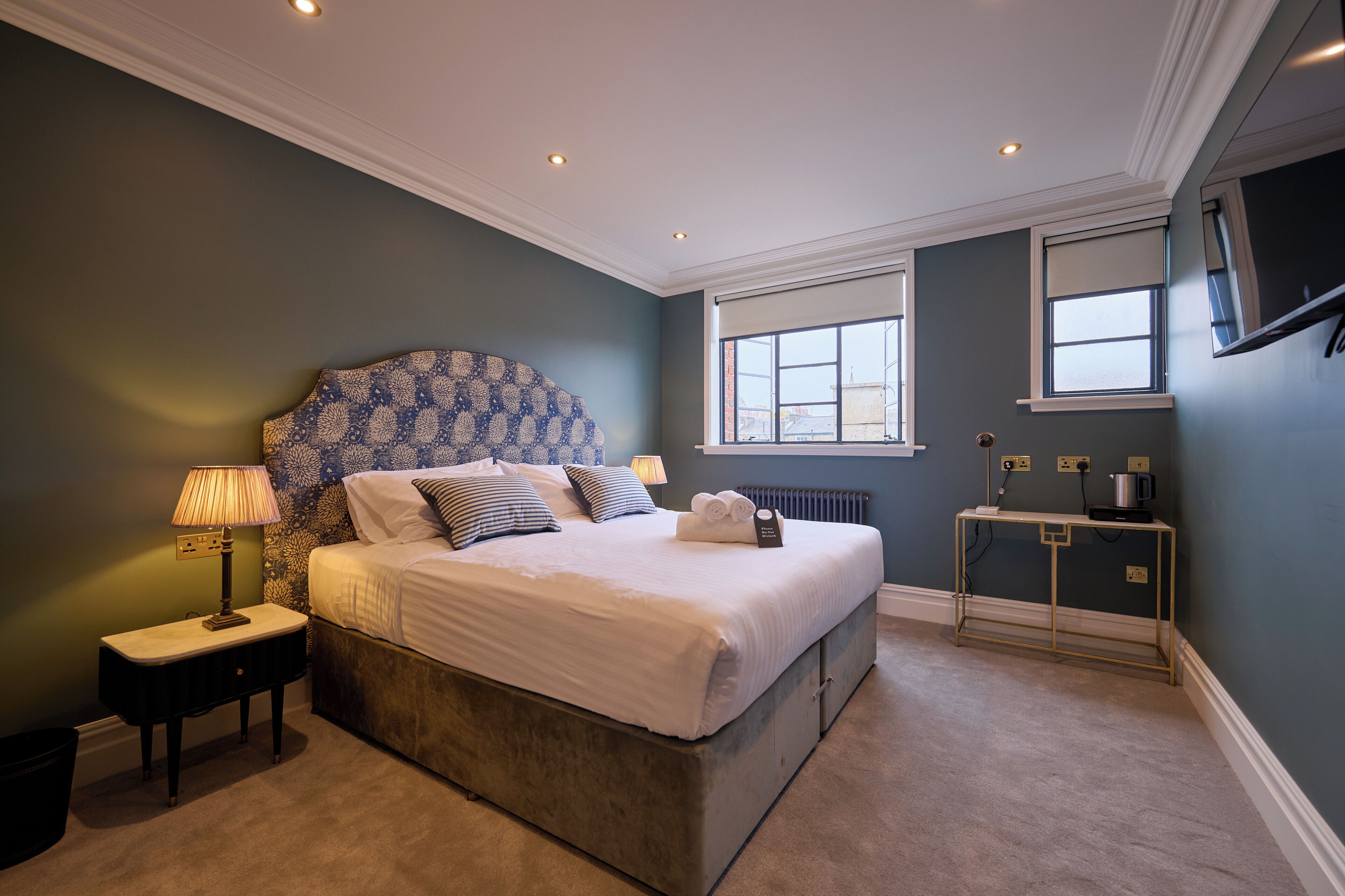 A bedroom with a large bed, a nightstand, a lamp, a radiator, and a TV mounted on the wall.