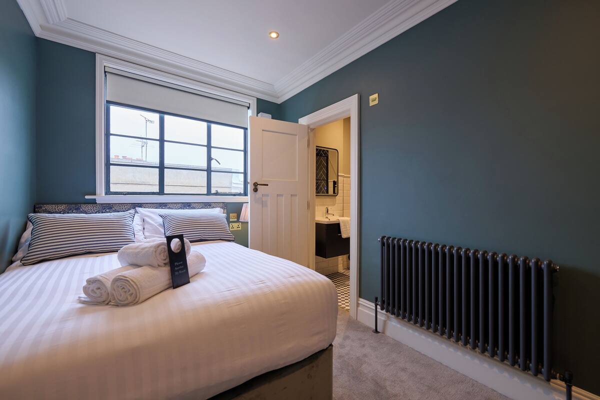 Una cama bien hecha con toallas enrolladas, una ventana con persianas, un radiador y un baño visible a través de una puerta abierta.