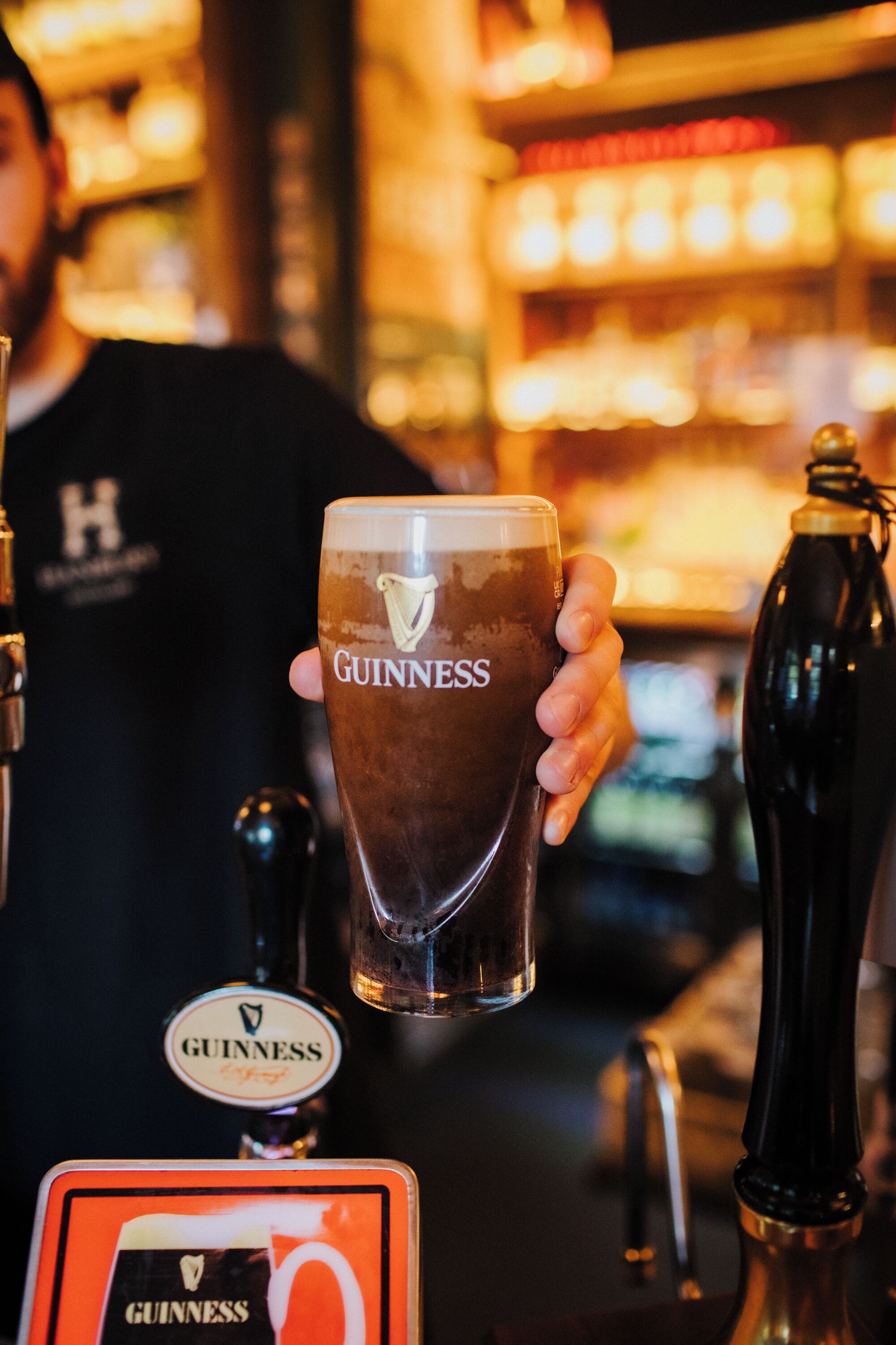 A person holding a Guinness glass at a bar with visible Guinness taps and a sign.