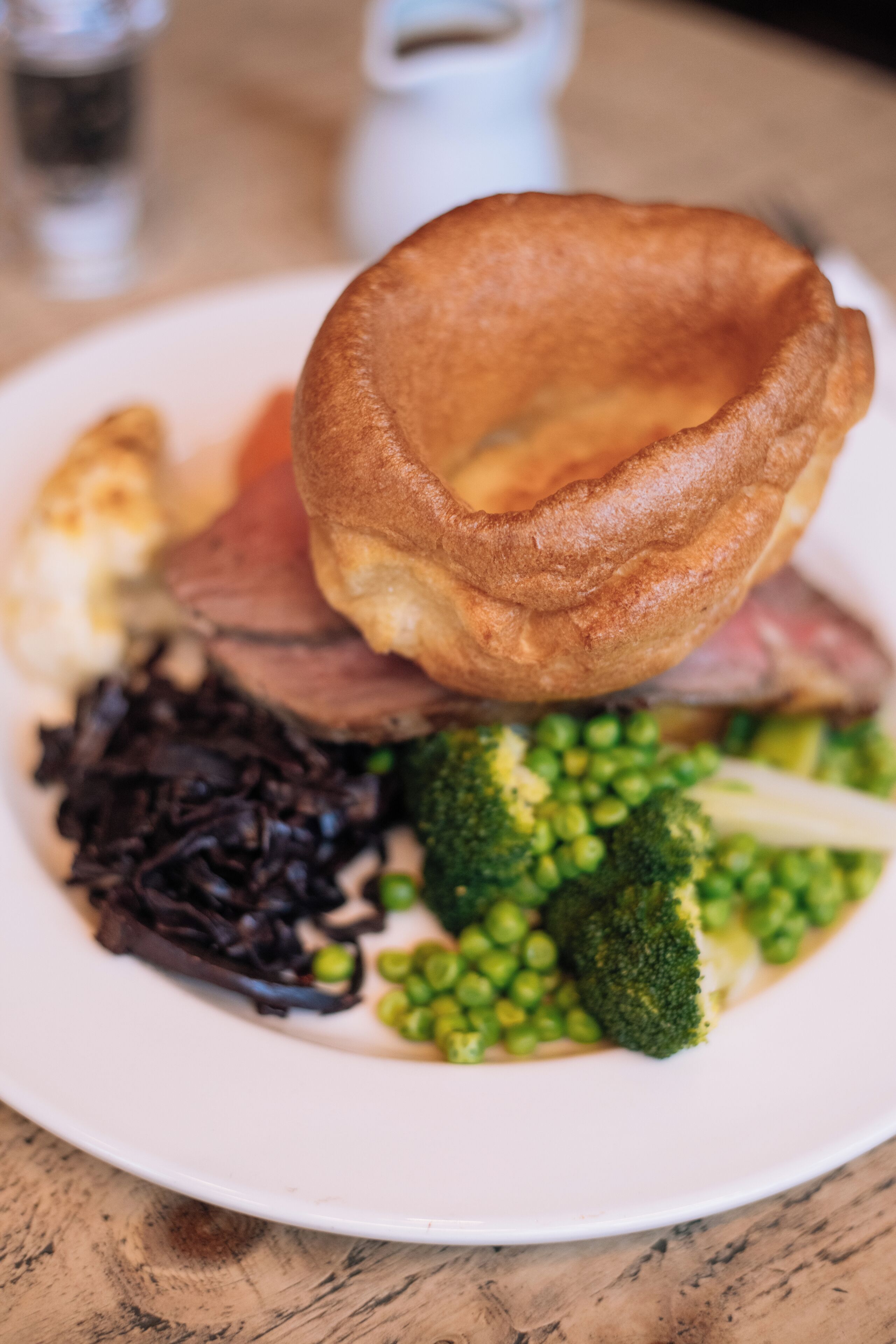 A plate of food with Yorkshire pudding, roast beef, broccoli, peas, and a side of what appears to be black pudding.