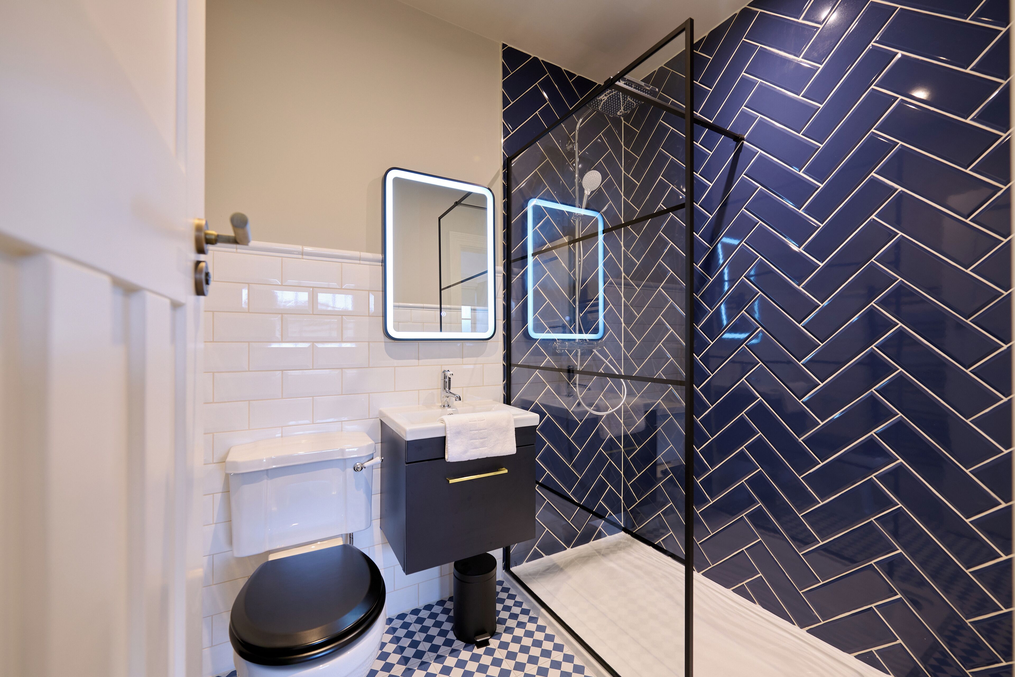 A modern bathroom with a black and white checkered floor, a black toilet, a white sink with a mirror, and a shower with a blue herringbone tile backsplash.