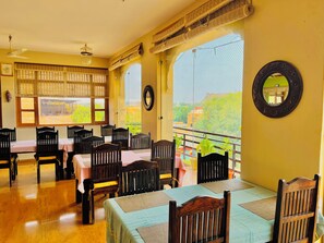 A dining area with tables and chairs, large windows with views, and a ceiling fan. - Jaisan Haveli (Jaisalmer)