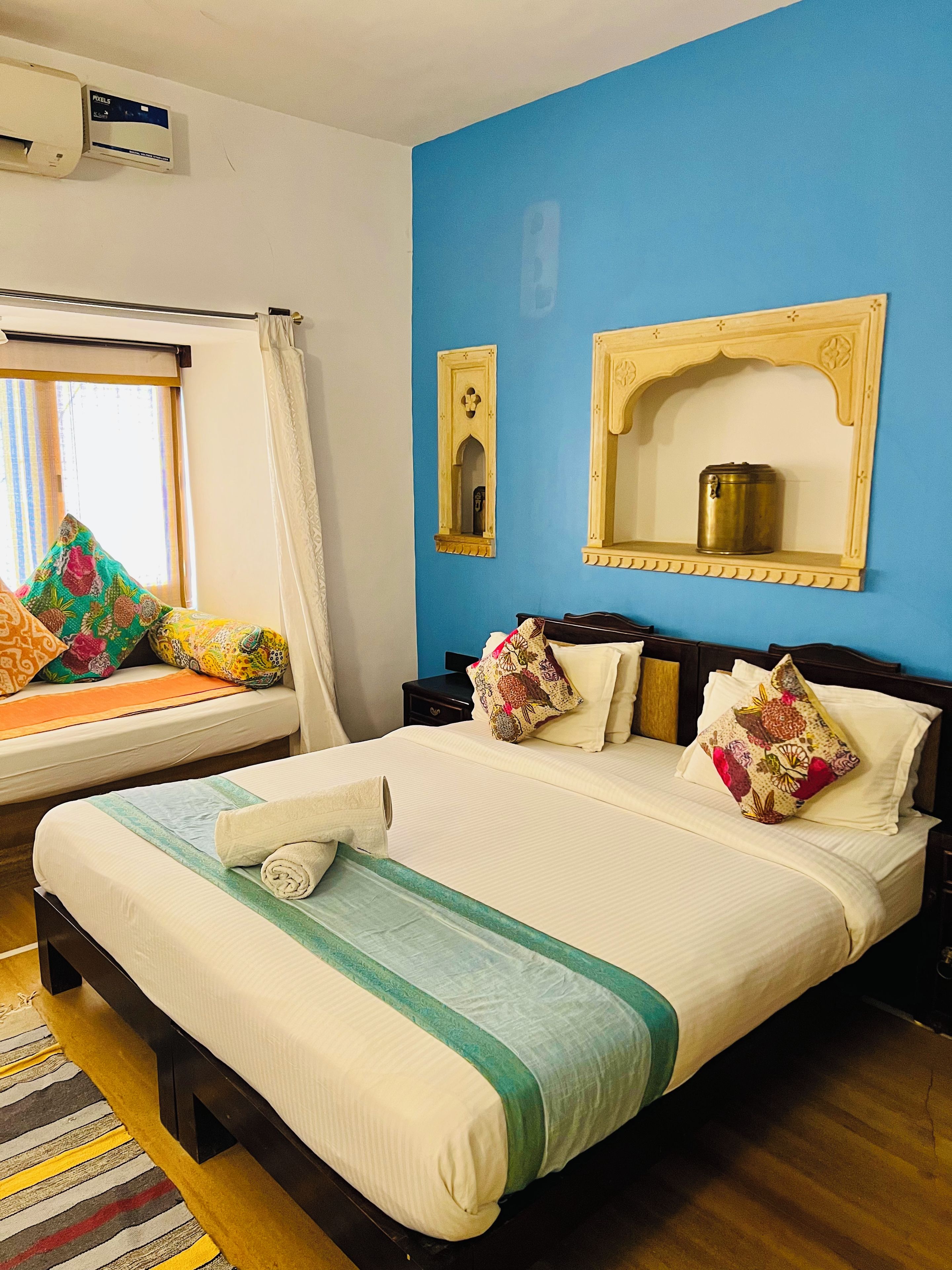 A hotel room with a large bed, a window with curtains, a wall-mounted air conditioner, and a small shelf with a container.