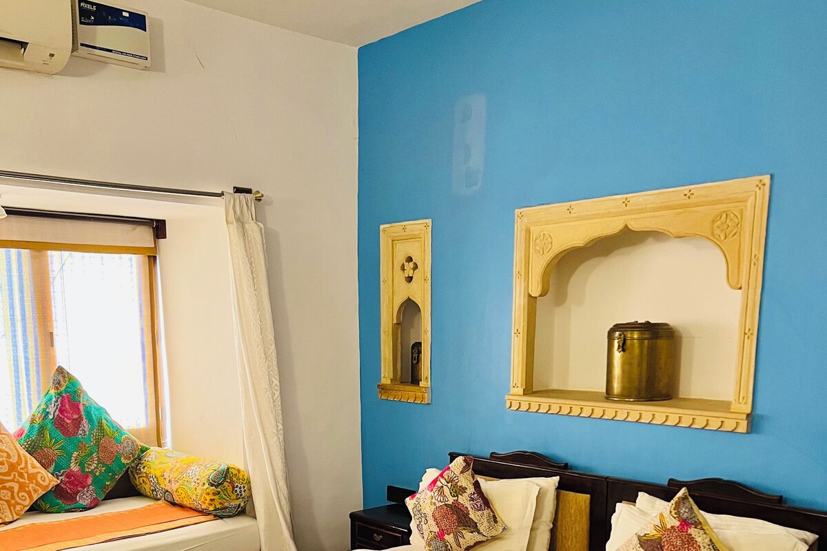 A hotel room with a large bed, a window with curtains, a wall-mounted air conditioner, and a small shelf with a container.