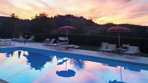 Piscina com espreguiçadeiras e guarda-sóis, rodeada por árvores e um céu ao pôr do sol.