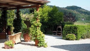 Uma área de estar coberta ao ar livre com pérgola, plantas em vasos e um forno rústico.