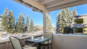 Un balcón con dos sillas y una mesa, con vistas a un paisaje nevado con altos árboles de hoja perenne y un edificio al fondo.