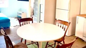 Un coin repas avec une table ronde et quatre chaises, une cuisine avec des meubles blancs et une porte-fenêtre donnant sur une autre pièce.